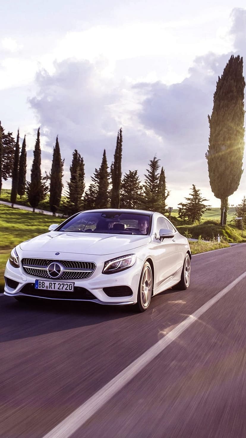 White Mercedes S-Class Coupe drives along a winding road lined with tall cypress trees under a dramatic cloudy sky.