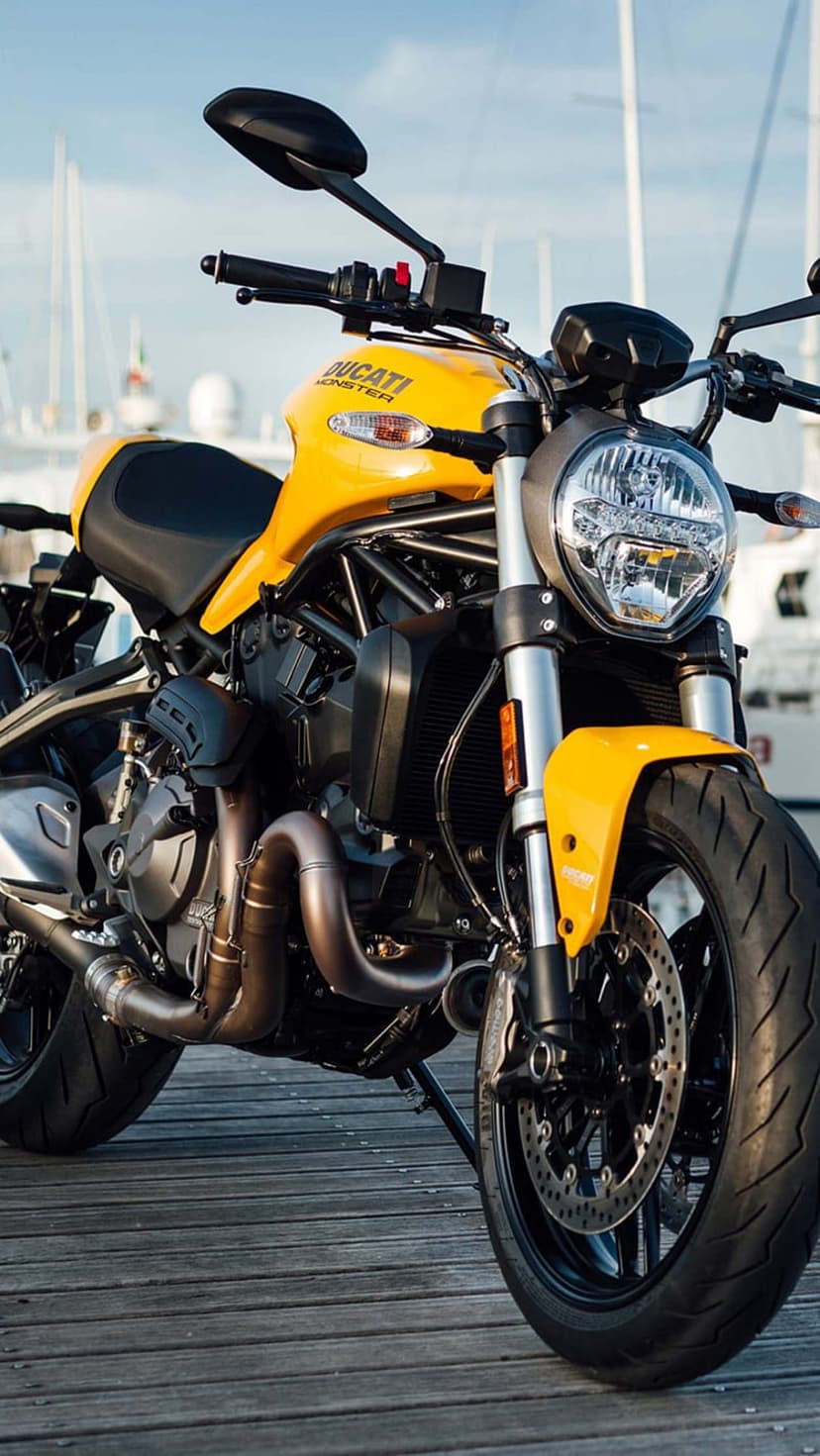 Yellow Ducati Monster motorcycle parked on a rustic wooden pier with blurred sailboats and blue sky background.