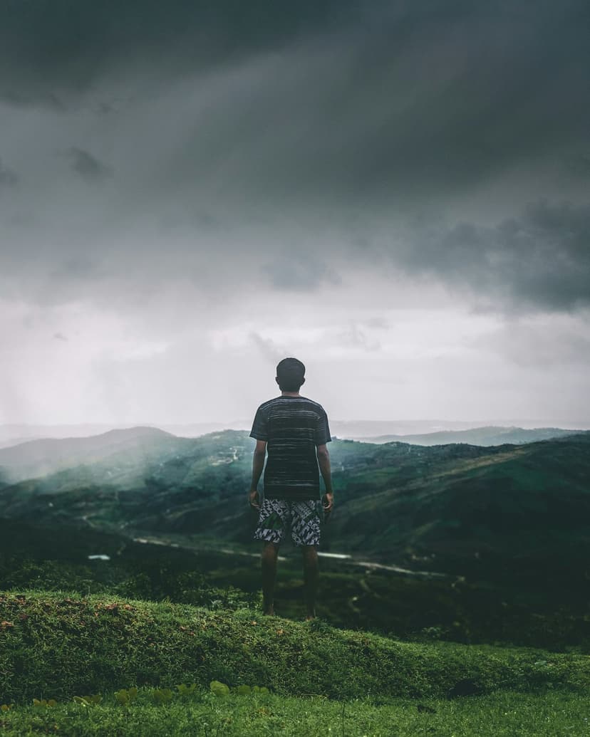 Man standing on a grassy ridge overlooks misty green mountains under a dark cloudy sky in a vertical view.
