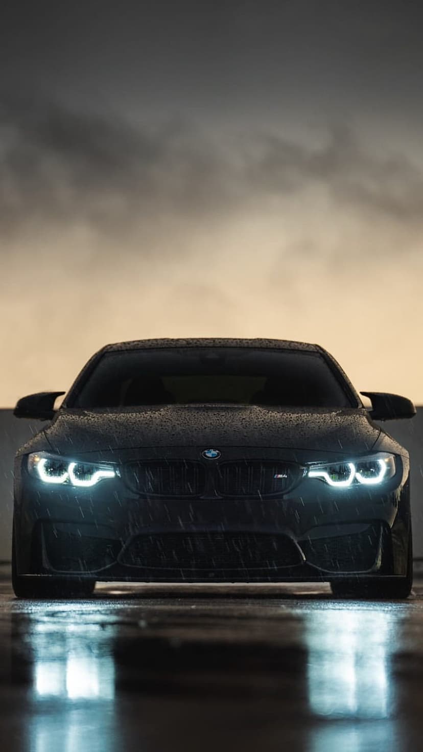 BMW M4 coupe with illuminated LED headlights parked on wet asphalt during a heavy rainstorm in a vertical view.