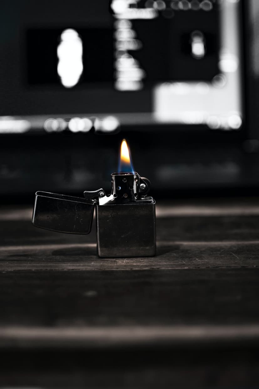 Silver metal lighter with an open lid and yellow flame resting on a dark brown wooden tabletop with bokeh lights.