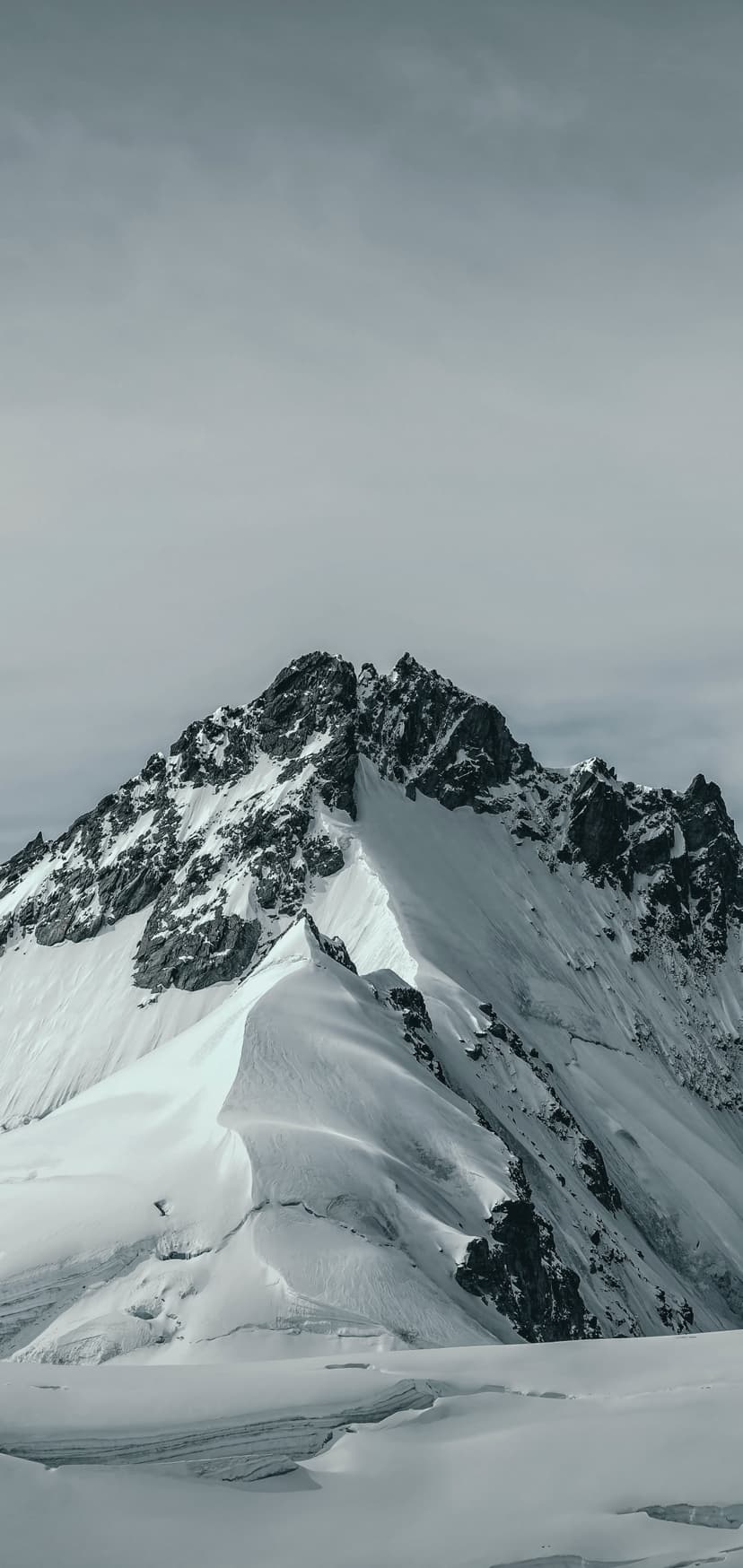 Sharp mountain summit covered in white snow against a dark grey overcast sky with rugged rock textures visible.
