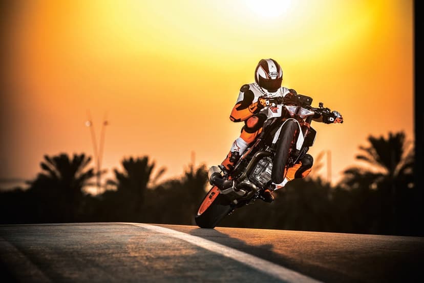 Motorcyclist doing a wheelie on an orange bike against a bright sunset with silhouetted palm trees.