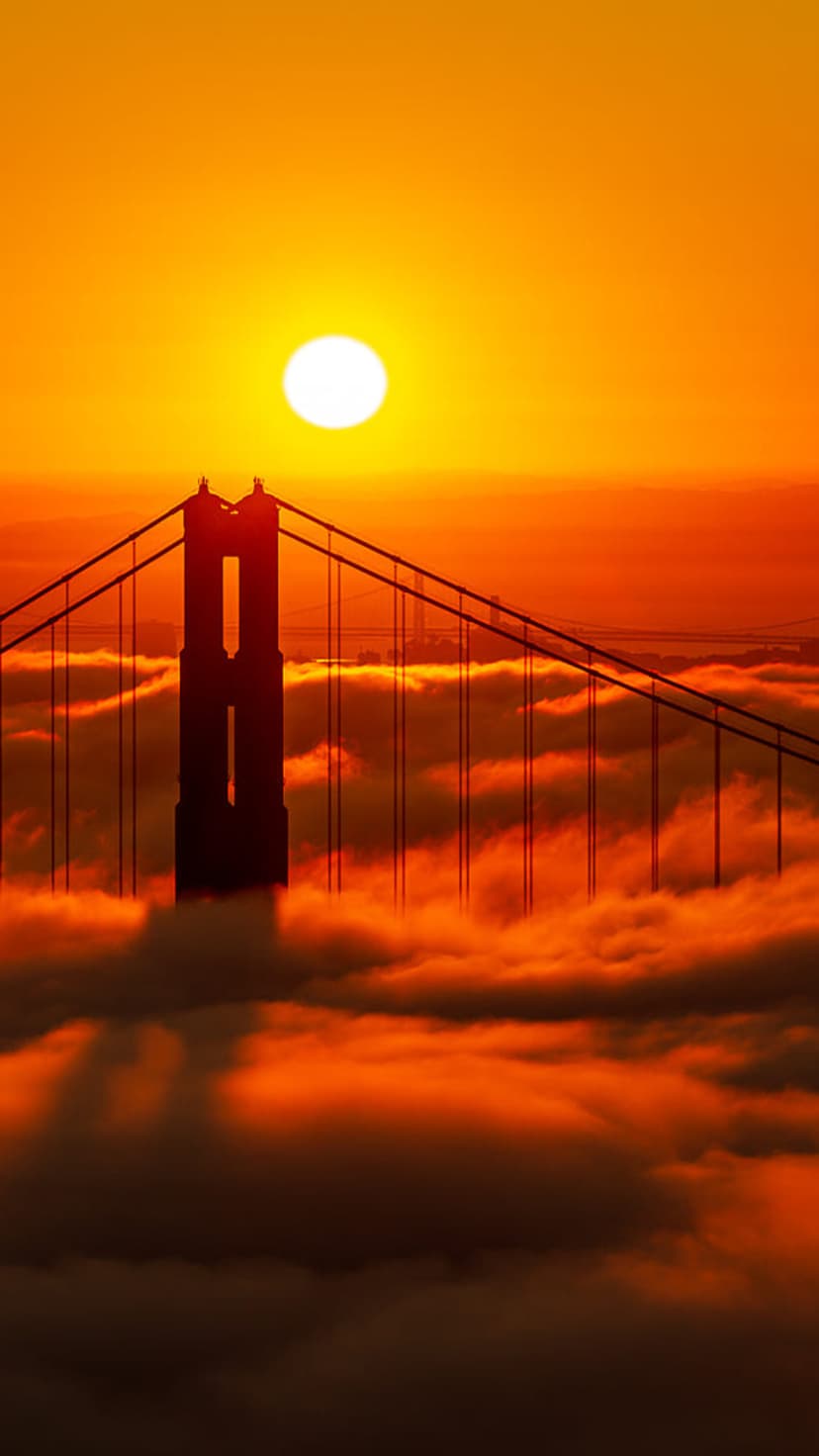 Golden Gate Bridge tower rising above a thick sea of fog under a brilliant orange and yellow sunset sky.