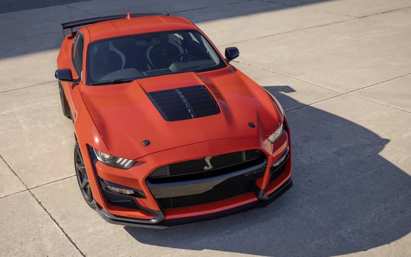 Orange Ford Mustang Shelby GT500 parked on concrete with long shadows and visible hood vents in sunlight.