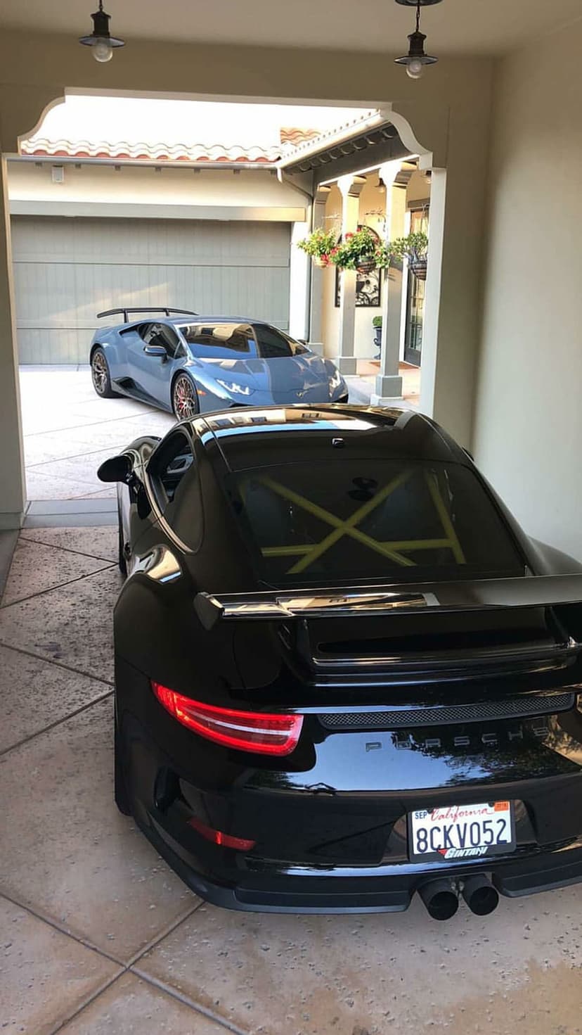Porsche GT3 in black with a roll cage and a blue Lamborghini parked in a garage entrance on a stone driveway.