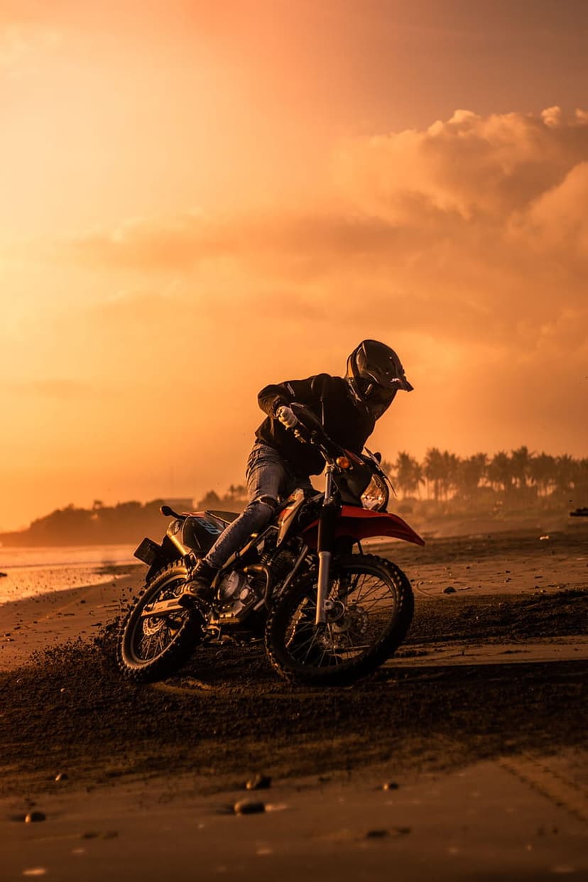 Motorcyclist riding a dirt bike along a sandy shoreline beneath a brilliant orange sunset with palm trees.