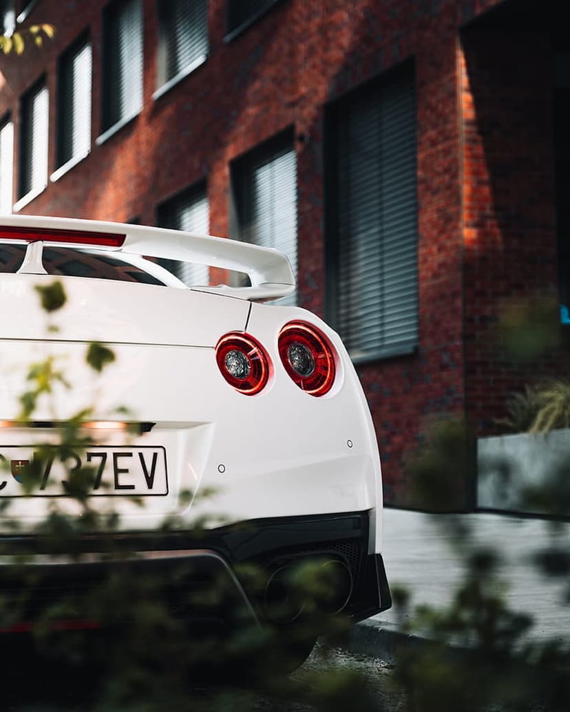 White Nissan GT-R rear view with round taillights and spoiler parked before a modern brown brick building facade.