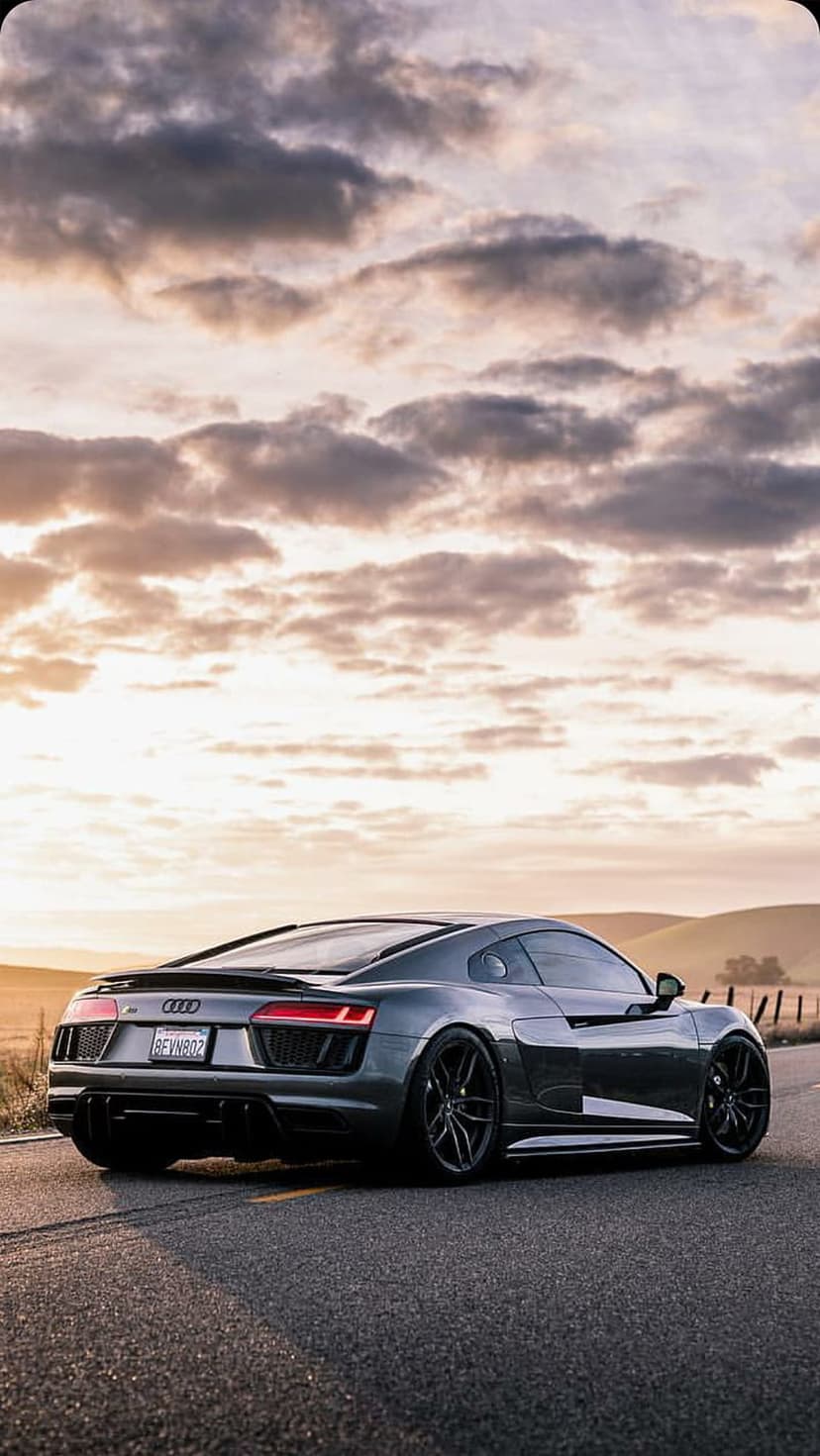 Audi R8 sports car drives on a scenic road against a backdrop of dramatic orange clouds and sunset lighting.