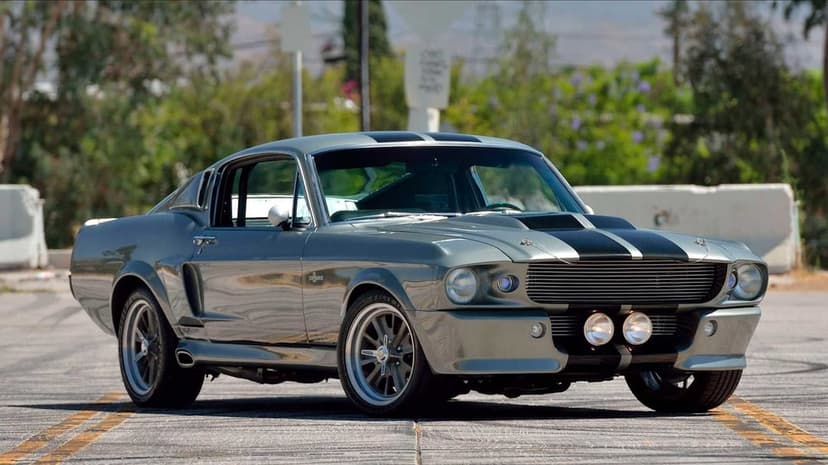Metallic grey 1967 Ford Mustang Eleanor with black stripes parked on asphalt under bright daylight.