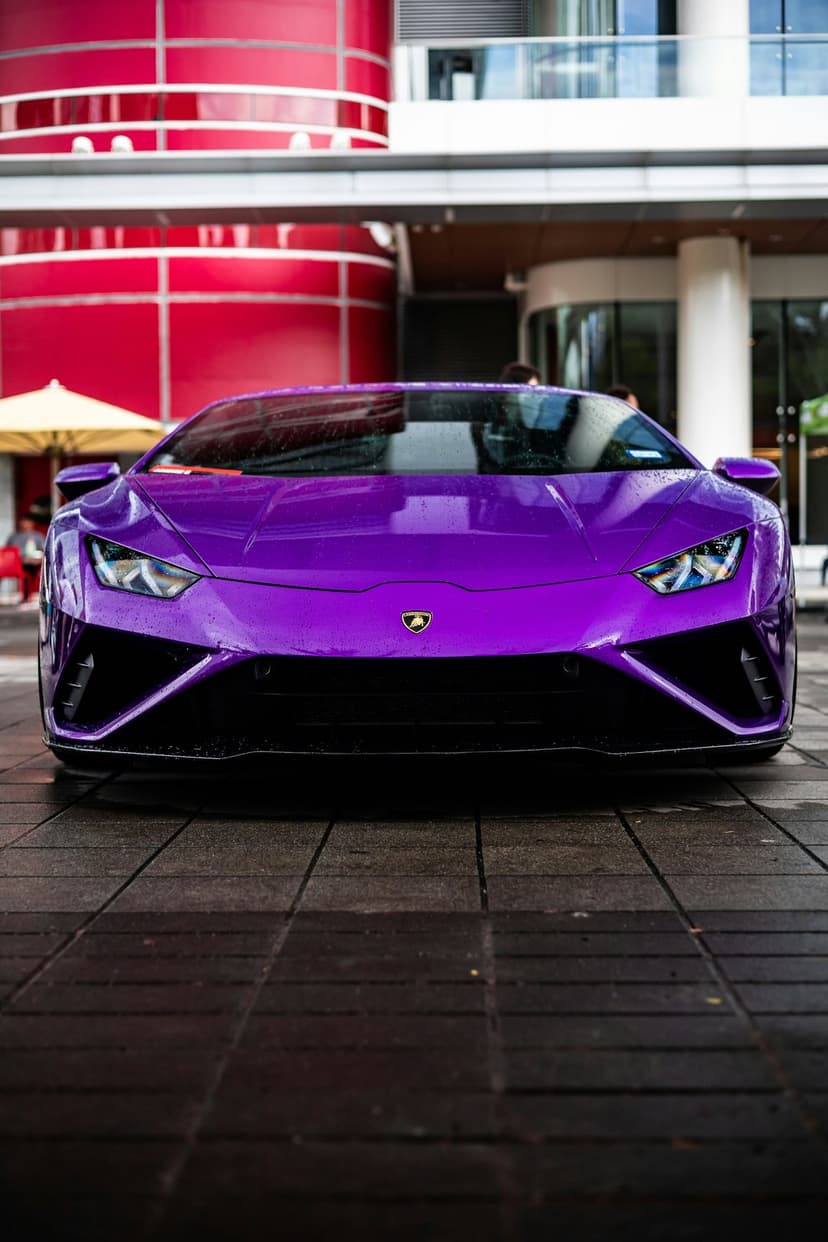 Purple Lamborghini Huracan parked on a wet surface with modern architecture and glowing headlights in view.