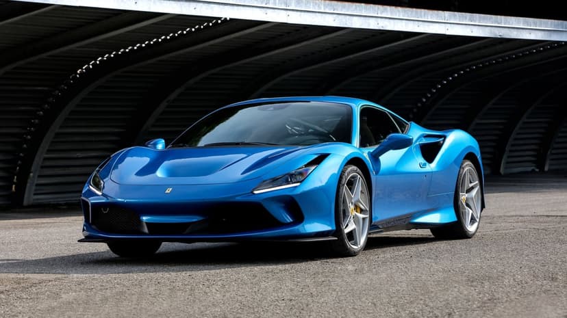 Electric blue Ferrari F8 Tributo parked on asphalt under a dark corrugated metal structure with side lighting.