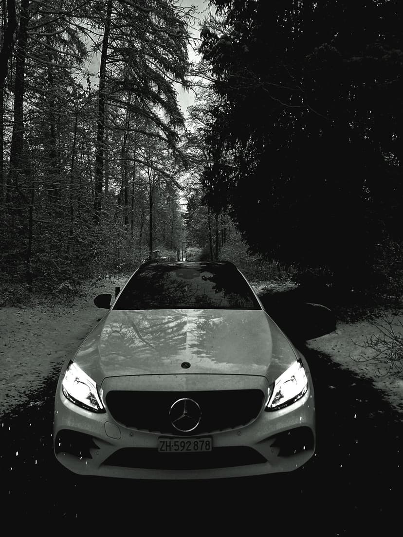 Mercedes-Benz sedan with glowing headlights parked on a snow-covered forest path between leafless winter trees.