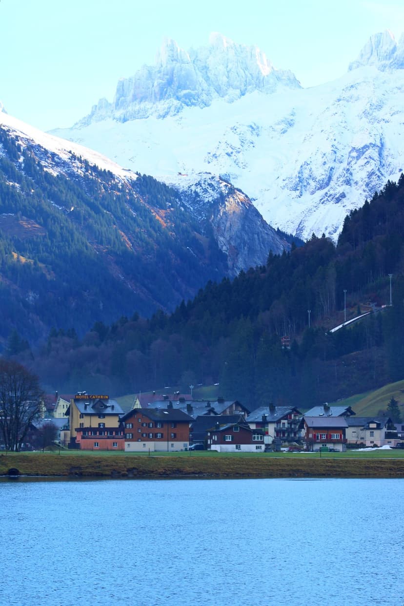 Colorful alpine houses sit along a calm blue lake with jagged snow-capped peaks rising in the background.