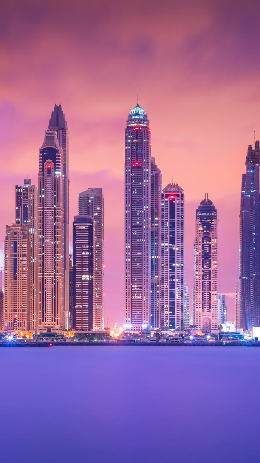 Glowing skyscrapers stand against a magenta and purple twilight sky, reflecting in a dark body of water below.