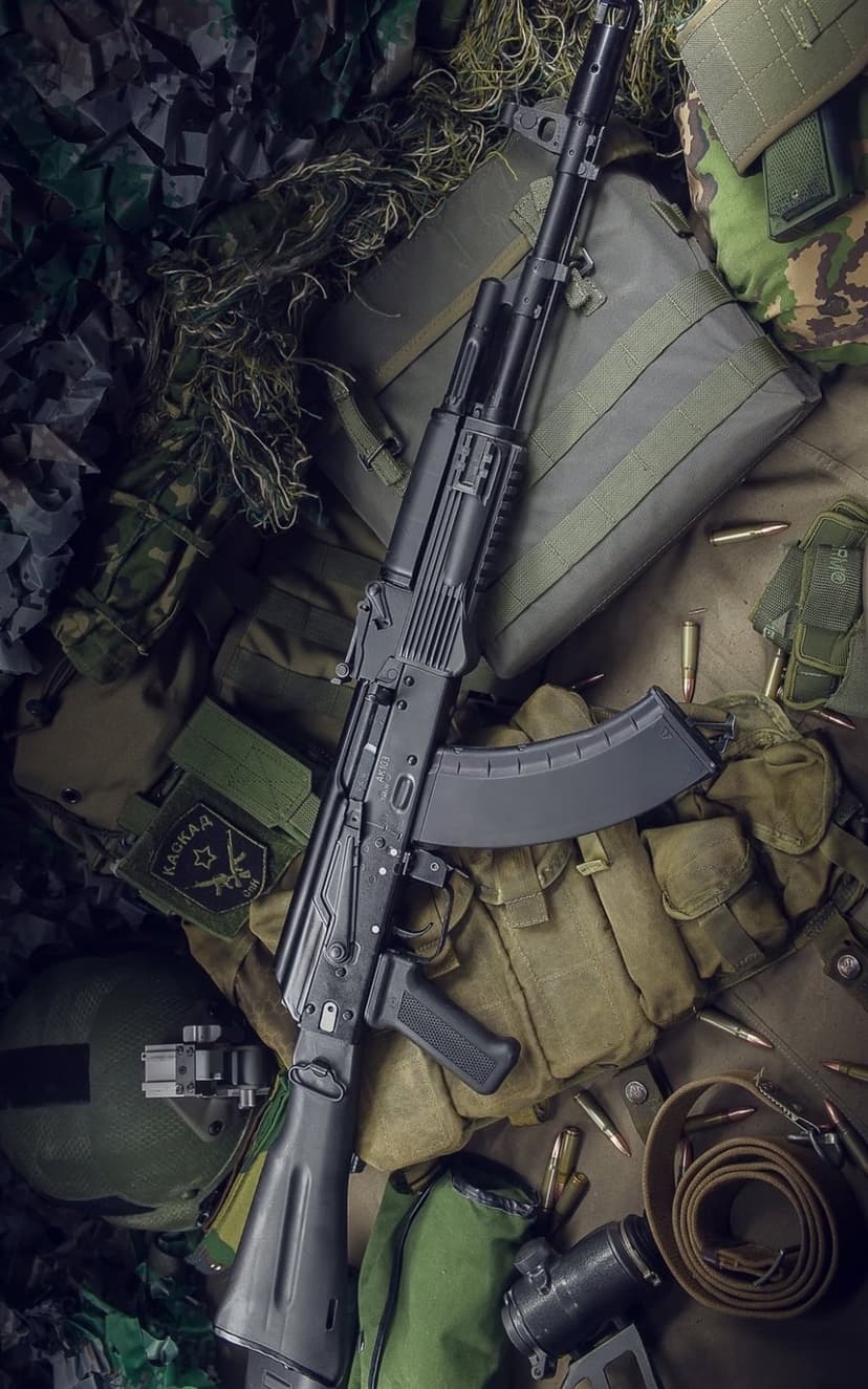 AK-47 rifle resting on camouflage netting next to a tactical helmet, ammunition rounds, and a green gear bag.