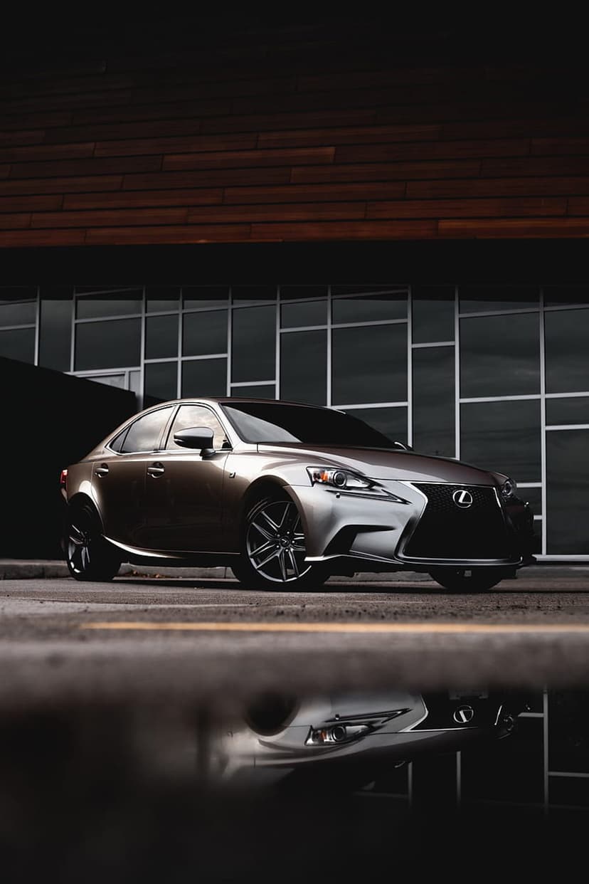 Lexus IS sedan parked near a wood-paneled building with its reflection visible in a wet puddle on the ground.
