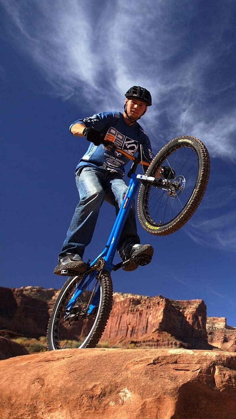 Mountain biker performs a wheelie on a blue bike atop red rock cliffs under a clear blue desert sky at midday.