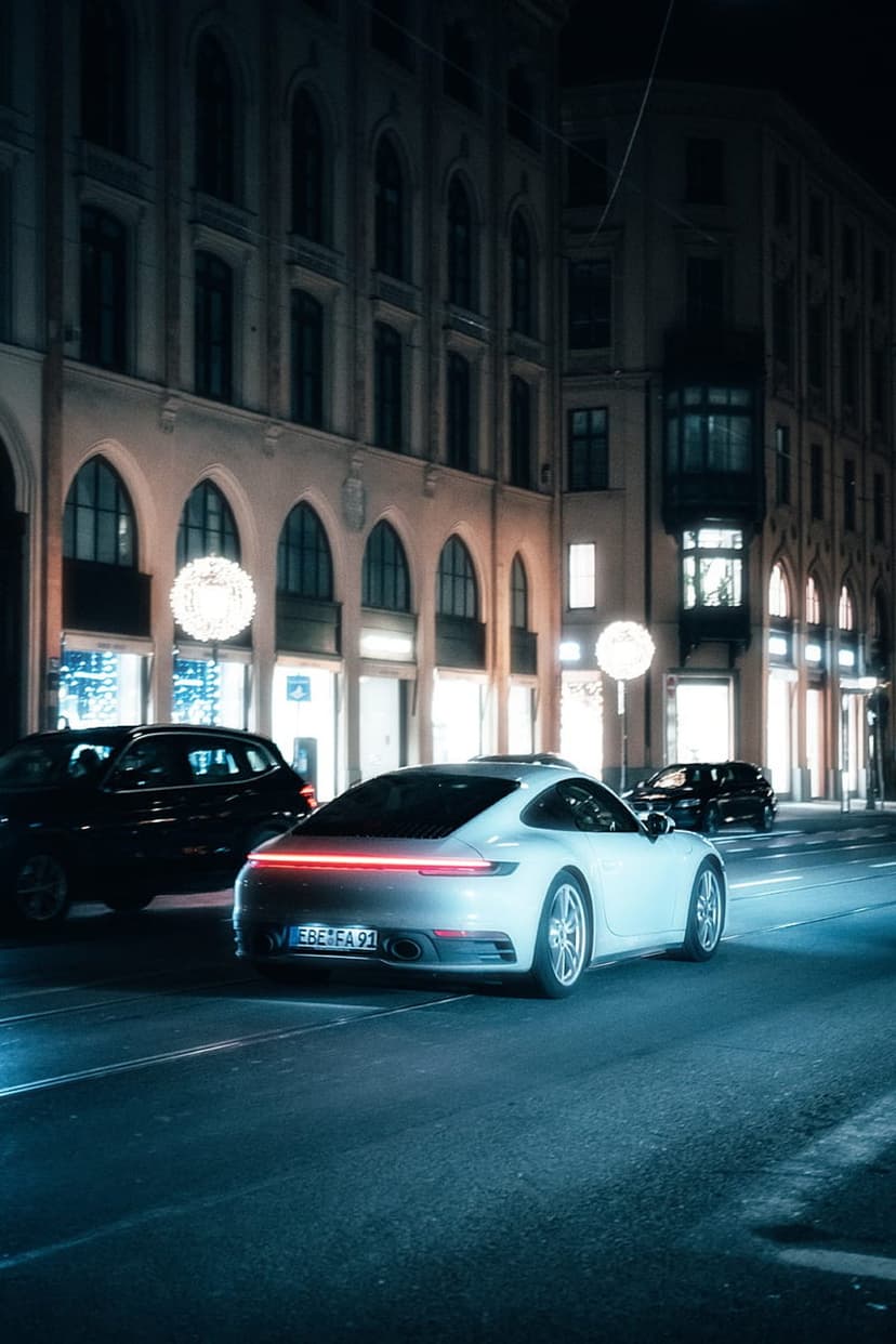 White Porsche 911 on a wet European street at night with red taillights reflecting on the dark ground.