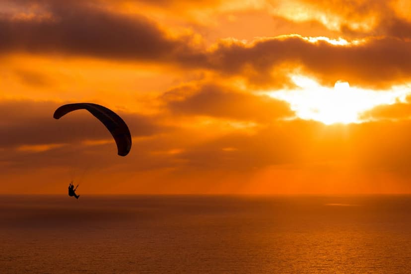 Paraglider silhouette glides above a glowing orange ocean under sunbeams breaking through dramatic evening clouds.