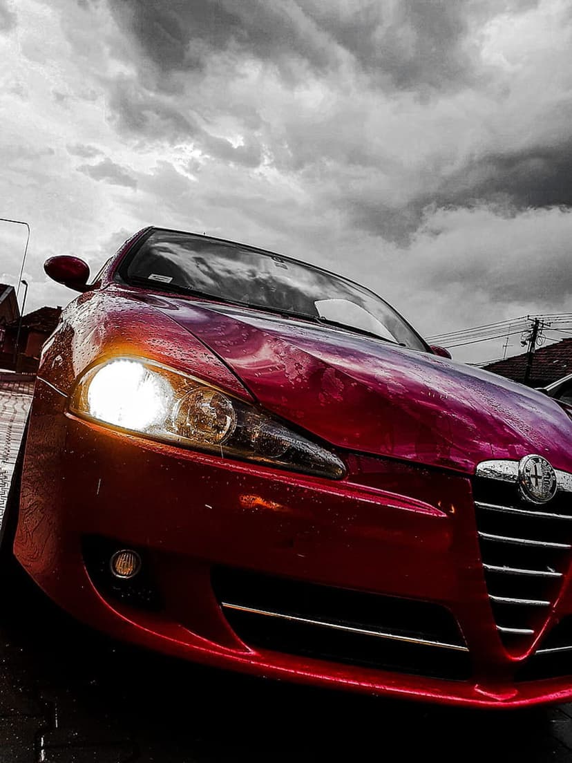 Red Alfa Romeo Brera with lit headlights parked on wet ground under a dark, cloudy, and dramatic stormy sky.