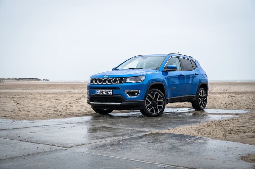 A vibrant blue Jeep Compass SUV parked on a wet paved ramp leading onto an expansive sandy beach under a cloudy sky.