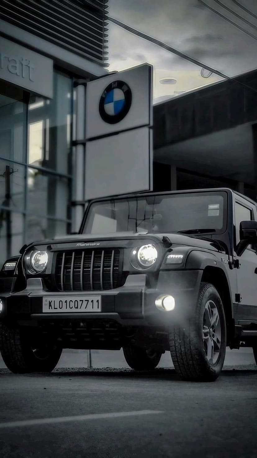 Black Mahindra Thar SUV with glowing headlights parked in front of a glass BMW dealership at night.