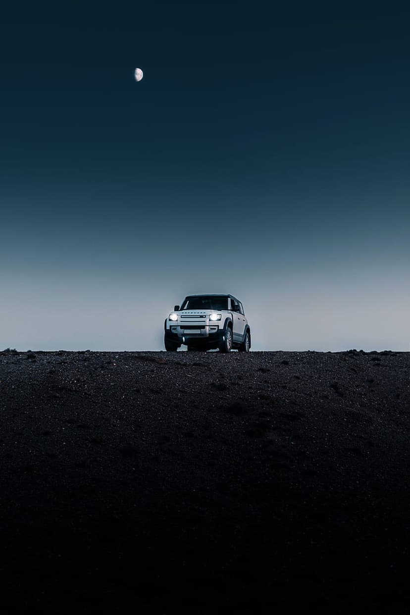 White Land Rover Defender SUV on dark textured terrain under a crescent moon in a deep blue night sky.