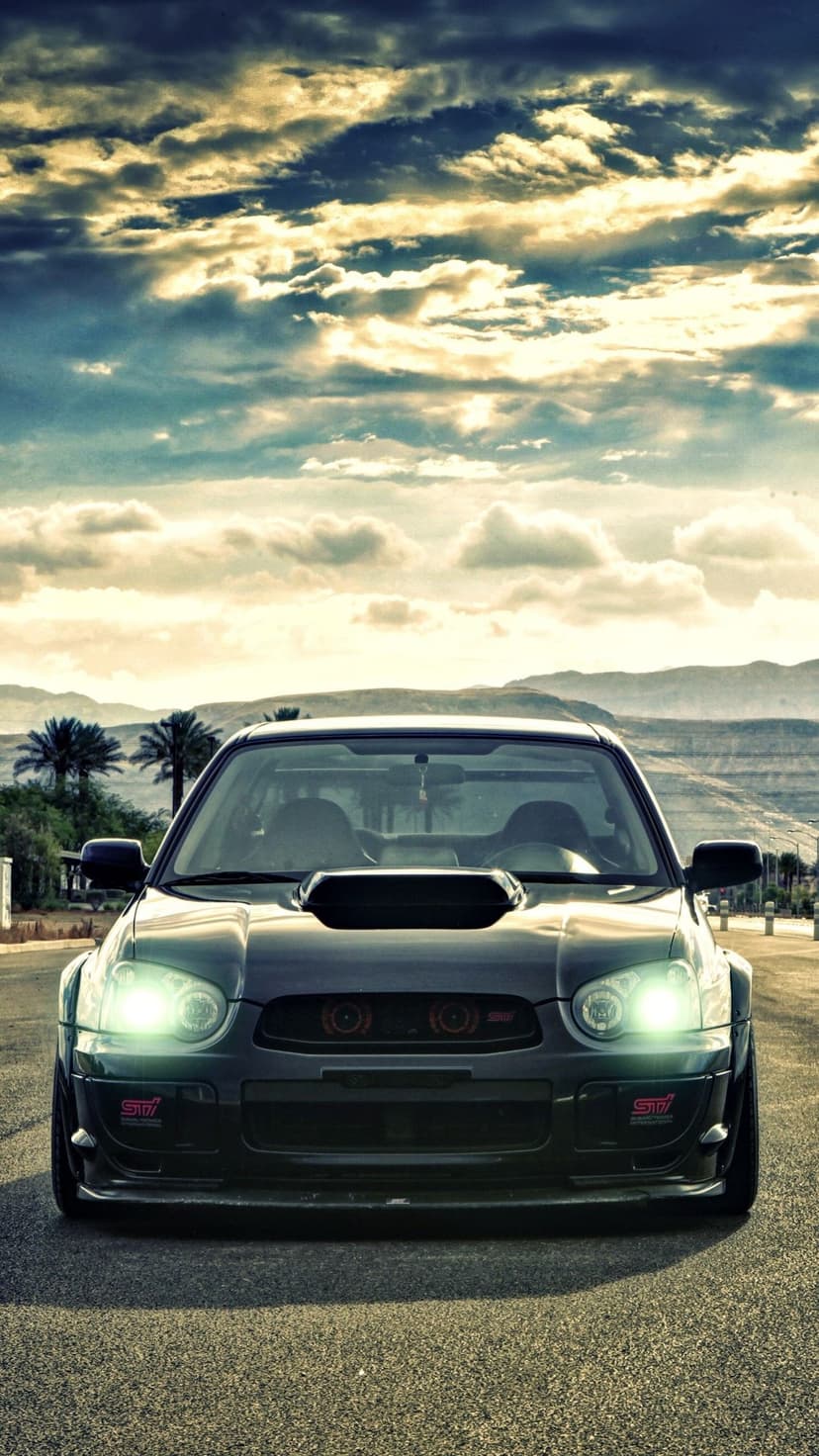 Dark Subaru WRX STI with glowing headlights on a desert road under a dramatic cloudy sky at sunset.