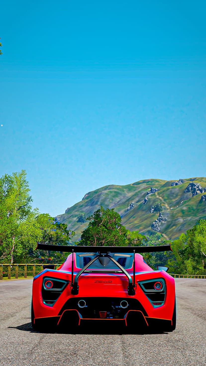 Red sports car with a large rear wing drives on a mountain road surrounded by green trees and rocky peaks.