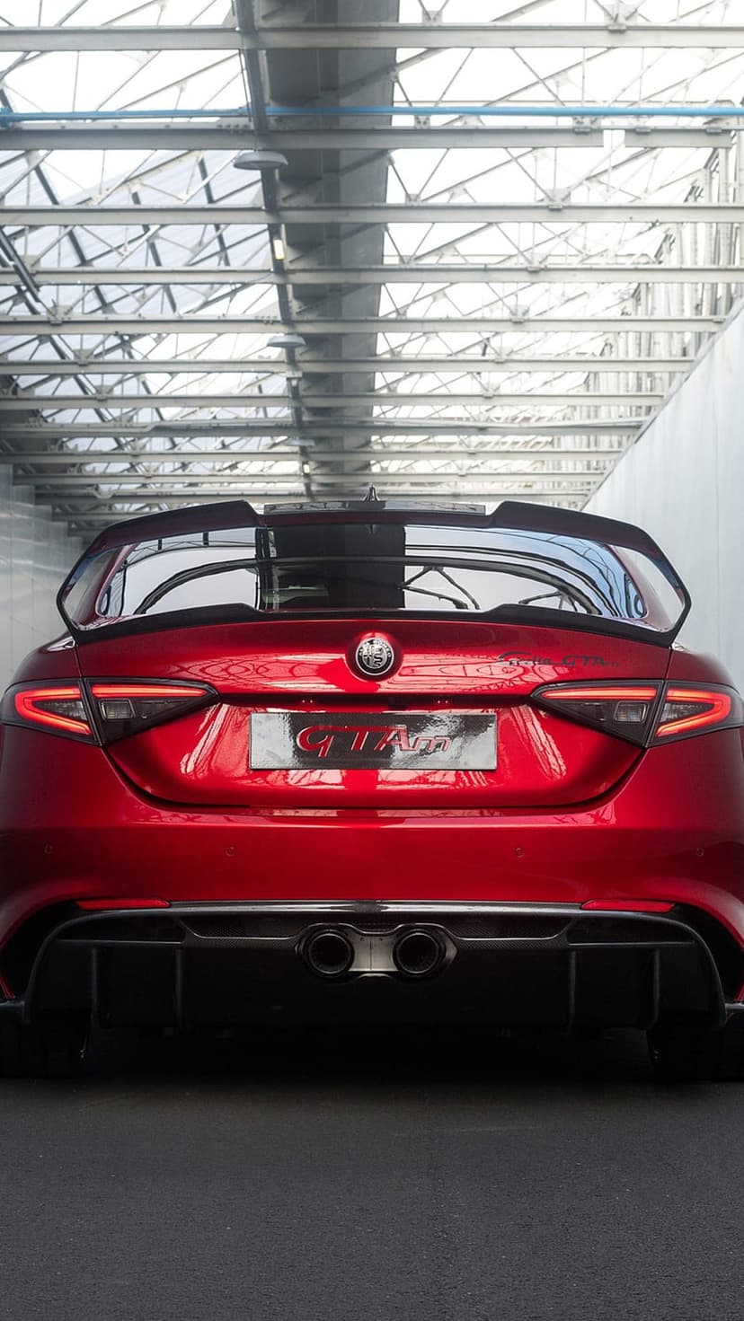 Red Alfa Romeo Giulia GTAm rear profile with a large wing and quad exhaust in a metal industrial garage.