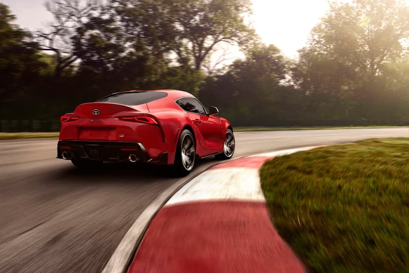 Red Toyota GR Supra driving fast on a racetrack curve with motion blur and forest background at sunset.