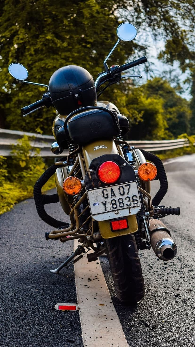 Royal Enfield motorcycle in desert sand color with a black helmet on the seat parked by a green forest road.