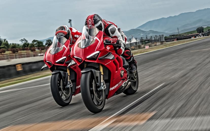 Two red Ducati sportbikes lean into a high-speed turn on a grey asphalt racetrack with distant mountains.