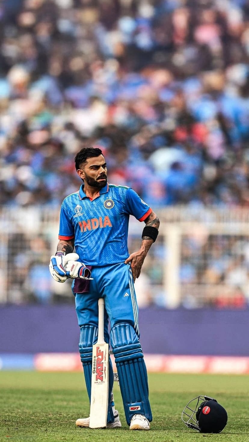 Virat Kohli holds a cricket bat while wearing a blue India jersey and pads on a green stadium grass field.