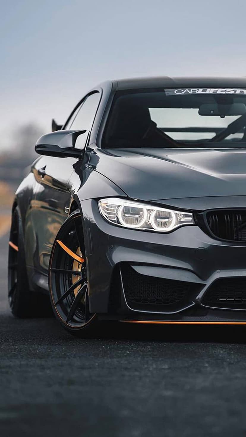 BMW M4 sports car in dark grey with orange accents and LED headlights parked on asphalt under a hazy sky.
