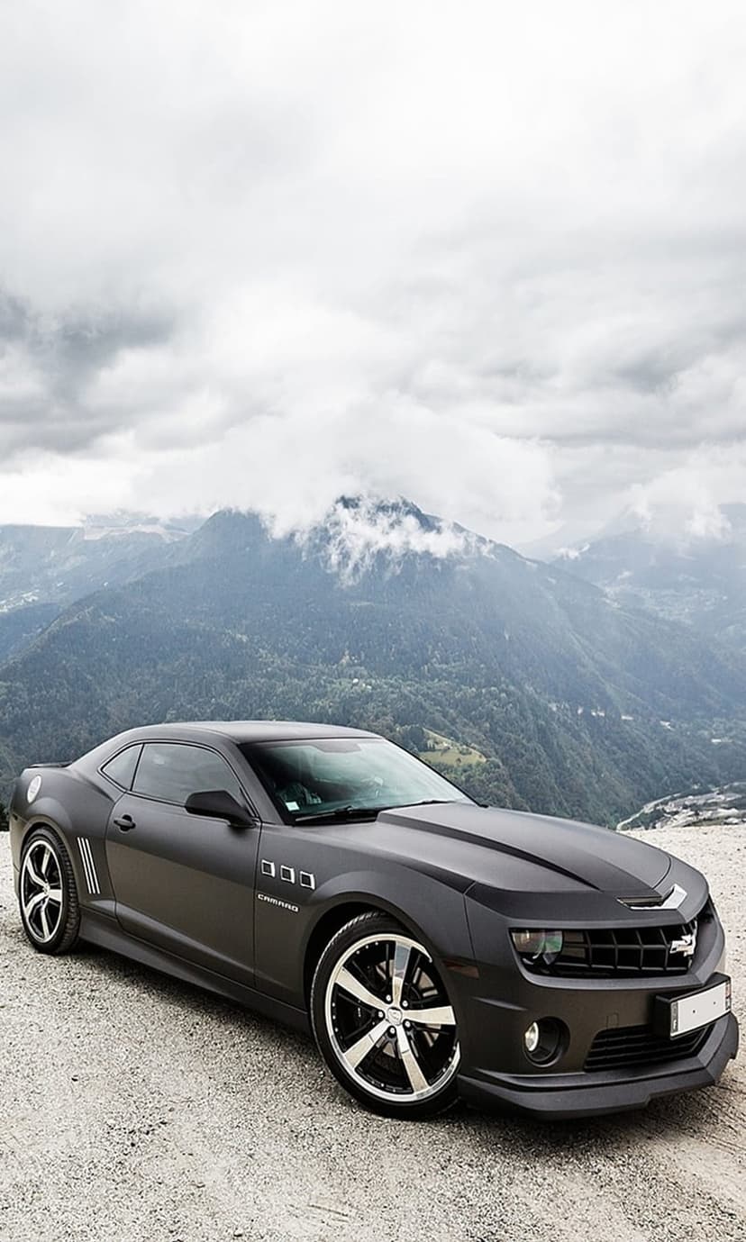 Matte black Chevrolet Camaro parked on a gravel mountain road under a cloudy sky with sharp custom rims visible.