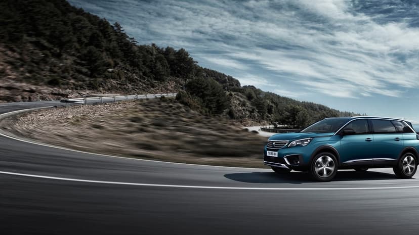 Teal Peugeot SUV driving around a sharp mountain road curve with motion blur and a cloudy sky background.