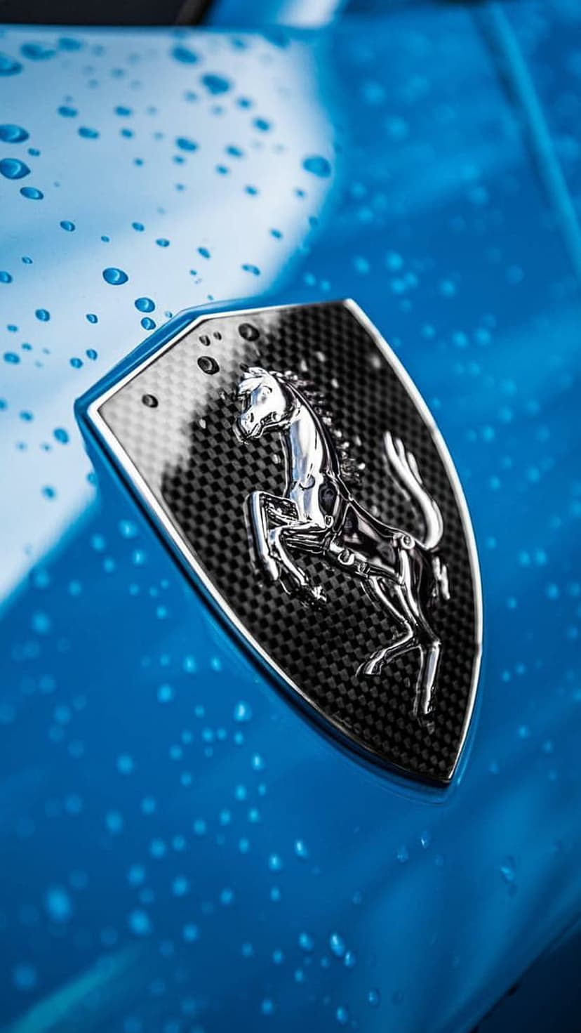 Silver Ferrari horse emblem on a carbon fiber badge fixed to a blue car hood covered in small water droplets.