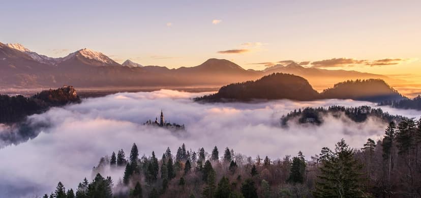 Bell tower church on a small island amidst violet valley fog with snowy mountains in the morning sunlight.