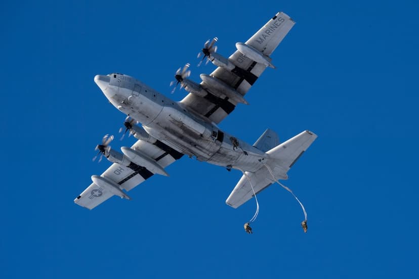 Marine paratroopers jump from a C-130 Hercules transport plane into a vast, sunlit clear blue sky from below.