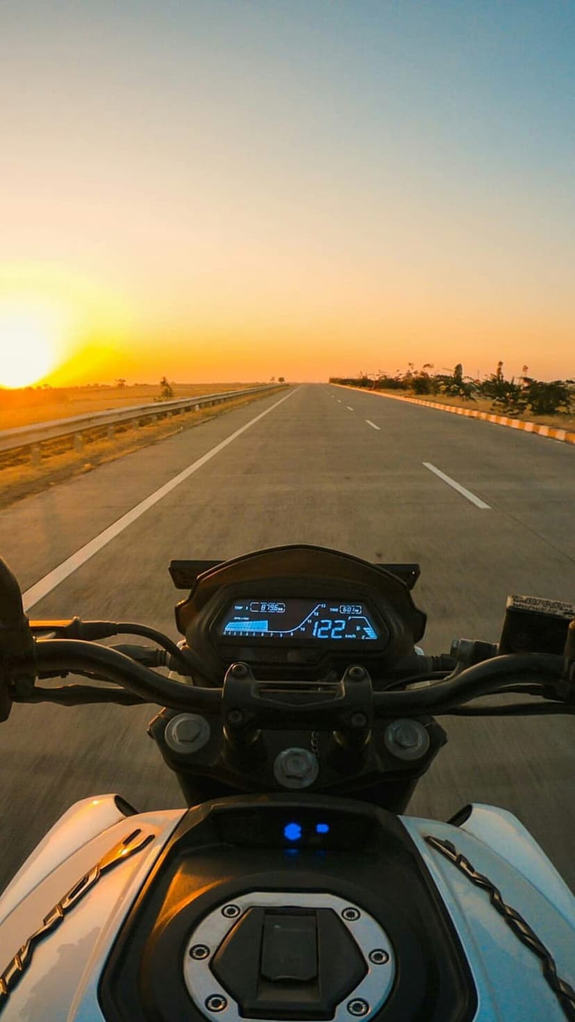 Motorcycle dashboard displays high speed on an empty highway at sunset with a glowing orange horizon and blue sky.