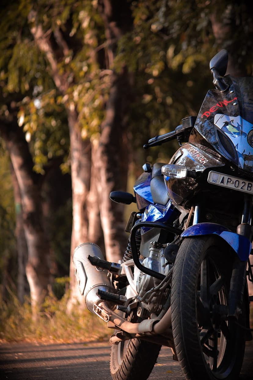 Blue and black motorcycle parked on a paved road with a blurred background of green trees and dappled sunlight.