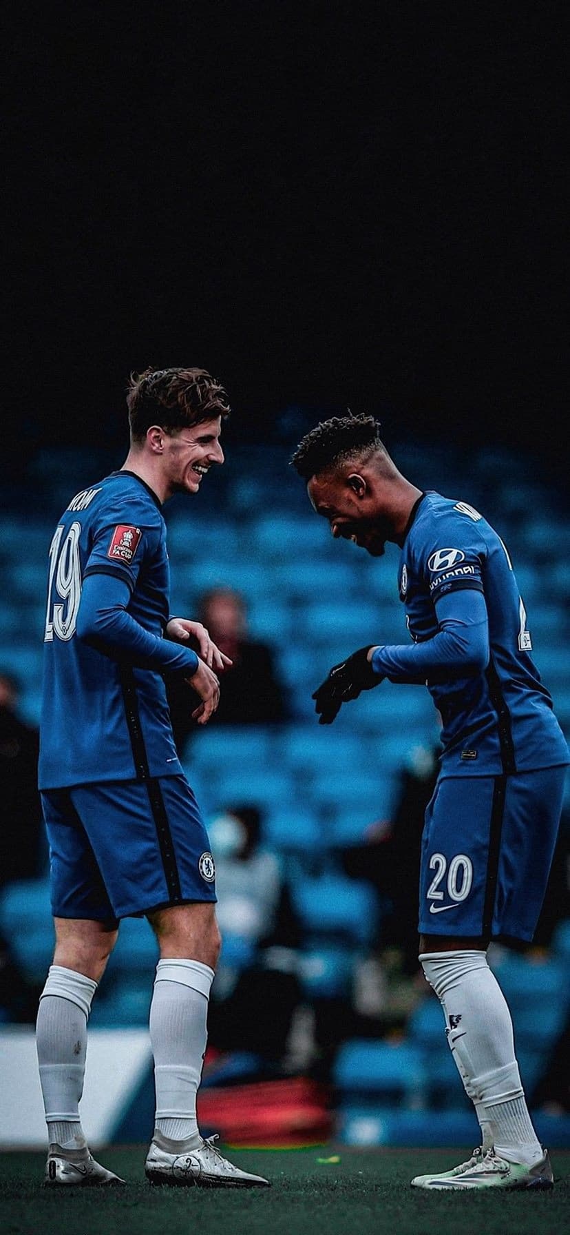Mason Mount and Callum Hudson-Odoi laugh together on a green football field wearing blue Chelsea FC kits.