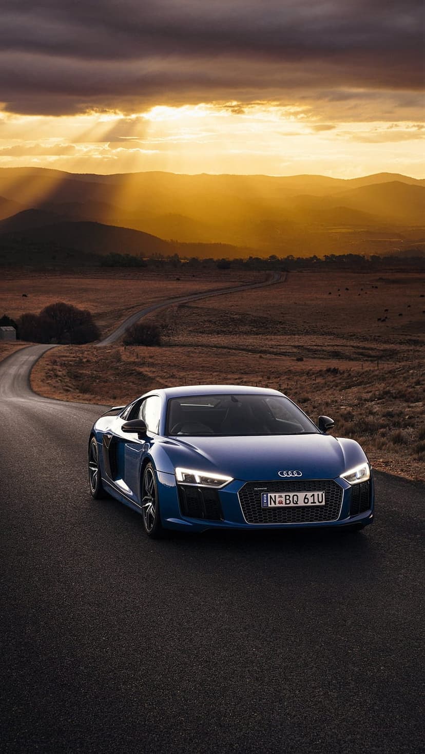 Blue Audi R8 sports car driving on a winding mountain road during a dramatic sunset with golden clouds and hills.