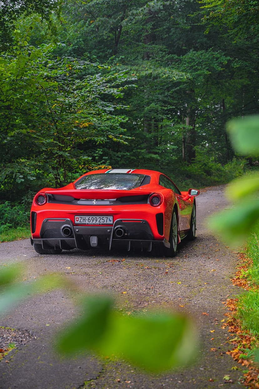 Red Ferrari 488 Pista seen from behind on a narrow road surrounded by dense green forest trees and foliage.