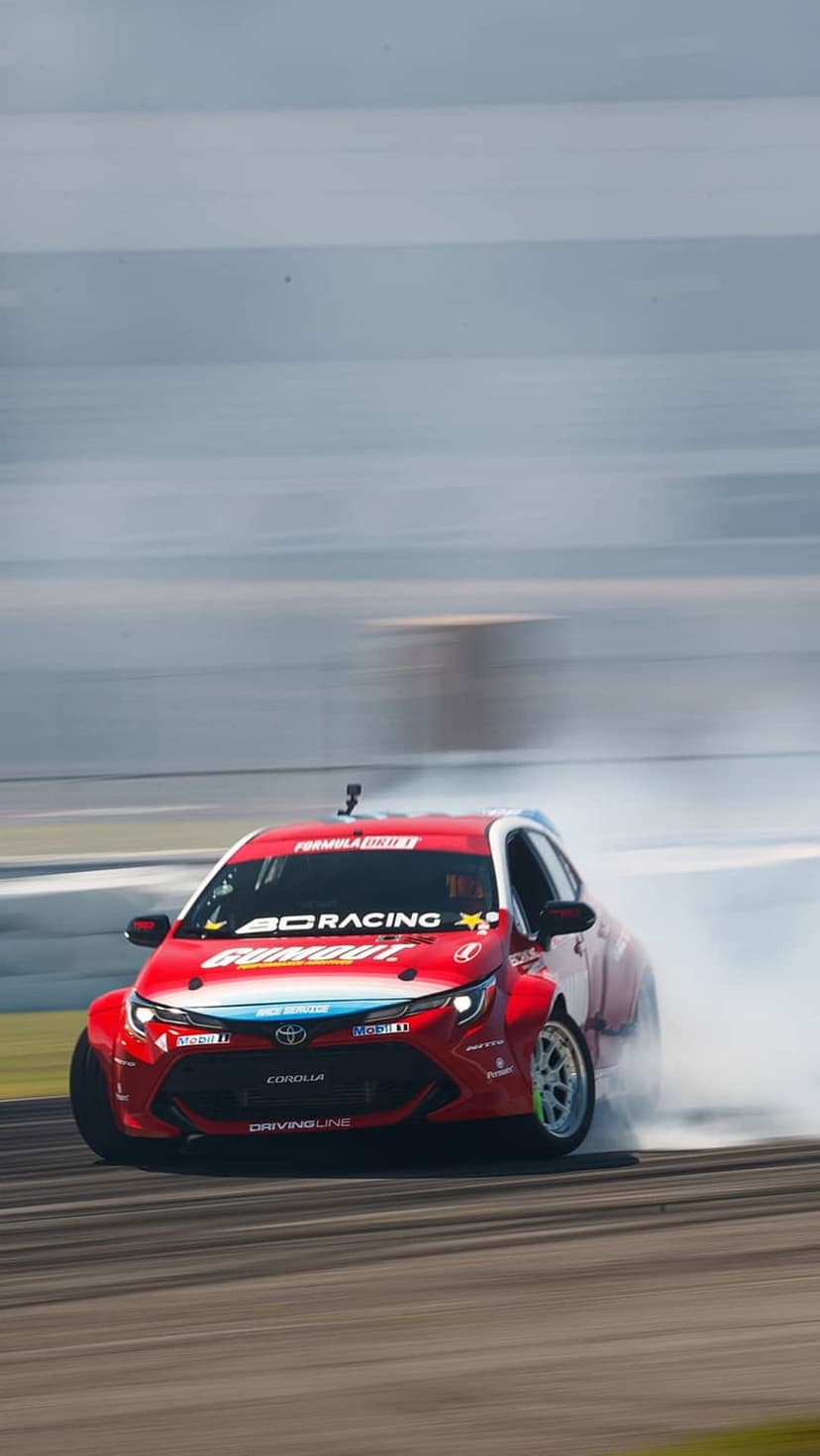 Red Toyota Corolla drifting through a turn on a racetrack with thick white tire smoke and motion-blurred background.