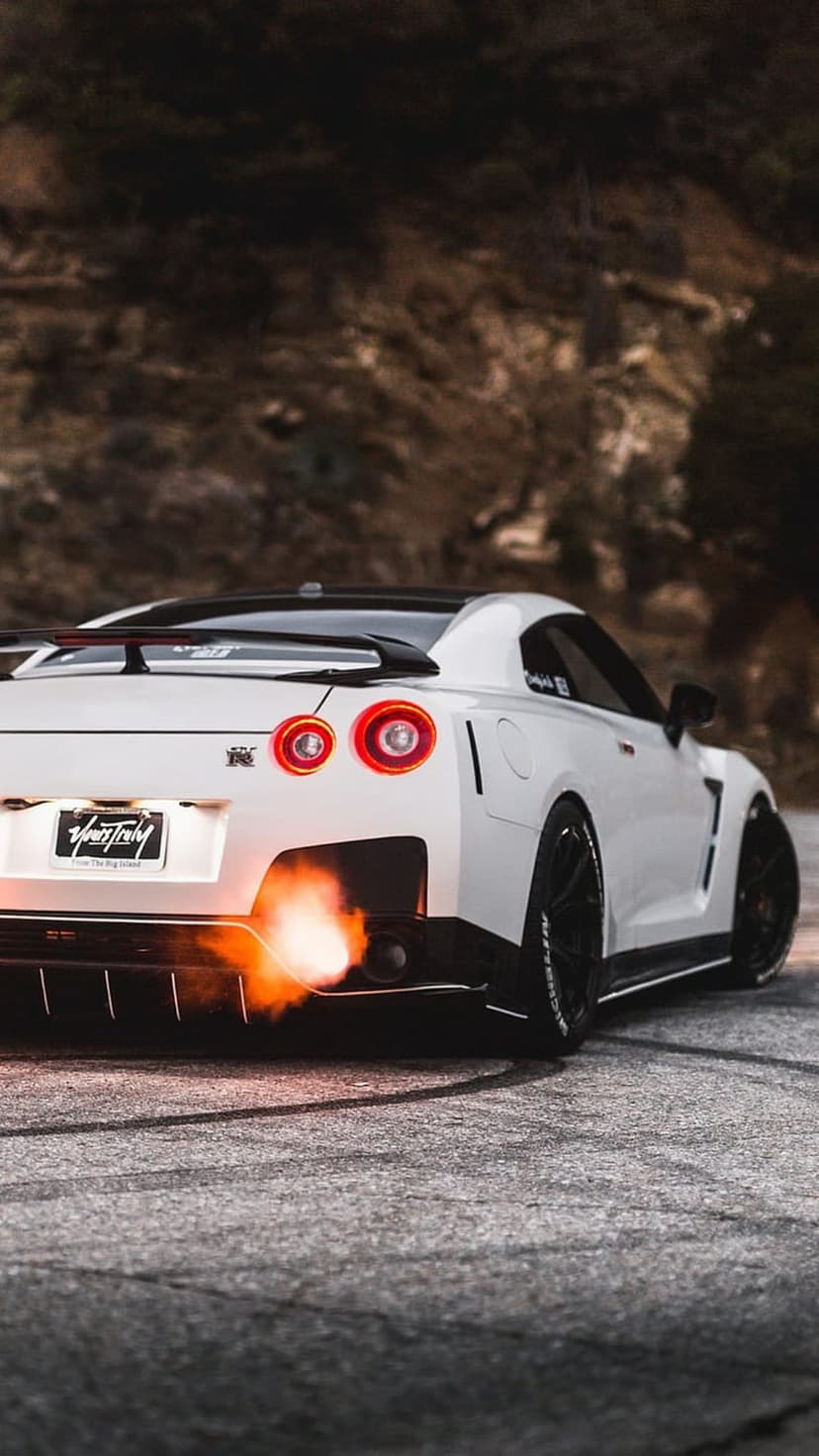 White Nissan GT-R supercar emitting orange exhaust flames on a paved road with blurred rocks and green trees.