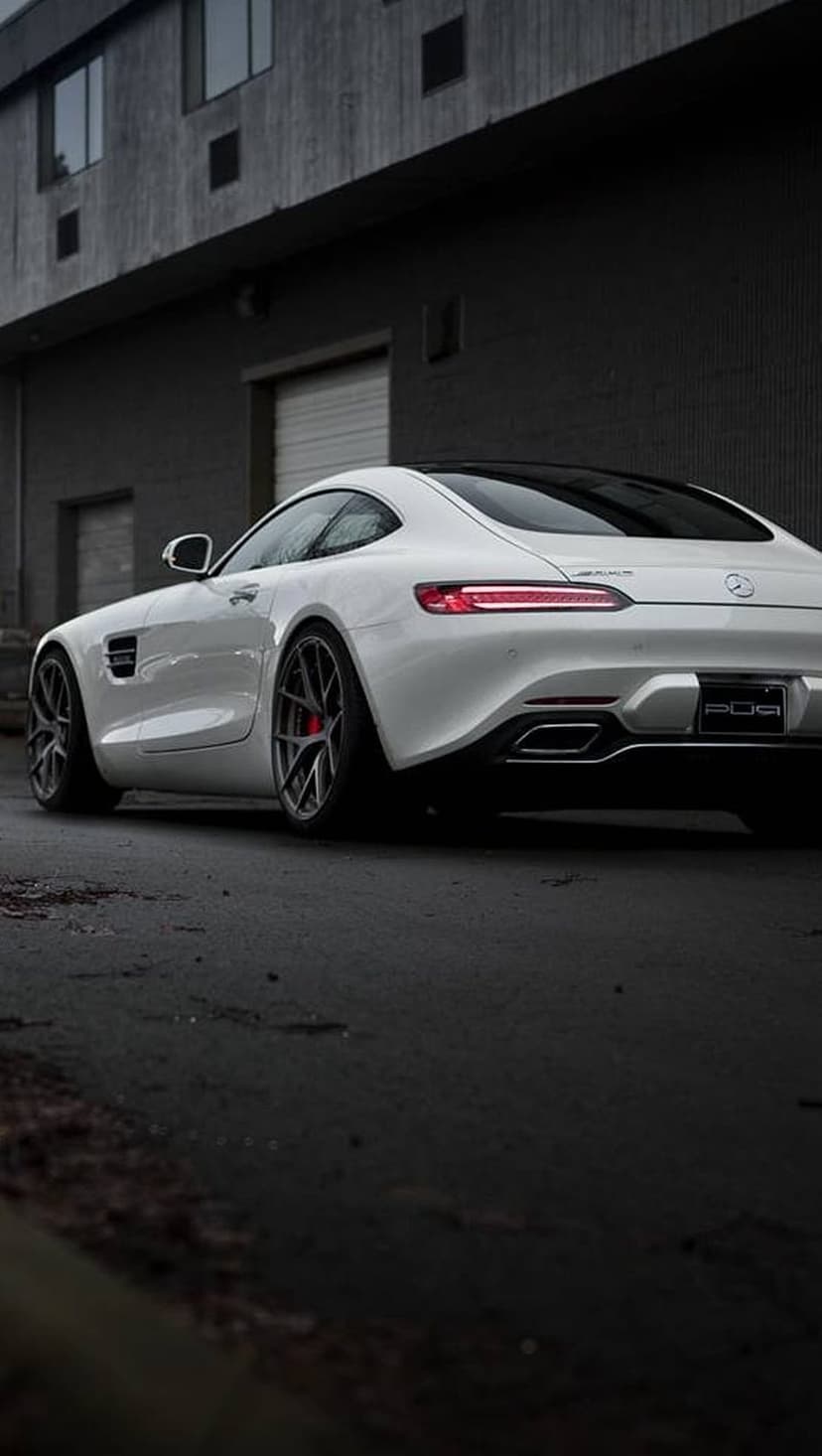 White Mercedes-AMG GT S rear view on wet asphalt beside a dark industrial building with illuminated windows.
