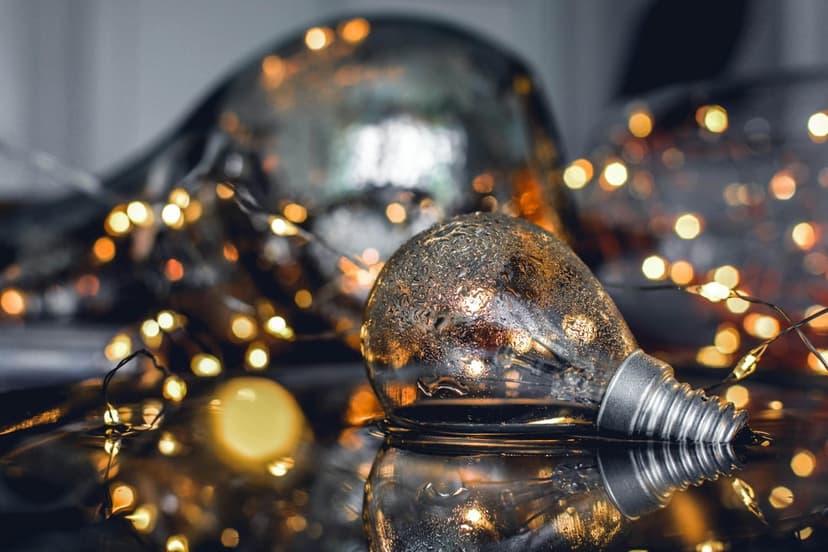 Glass Edison bulb ornament on a reflective surface surrounded by glowing golden fairy lights and blurred bokeh.